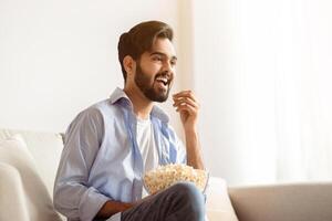 Cheerful Arab Guy Watching Tv And Eating Popcorn At Home, Relaxing On Couch In Living Room, Cheerful Eastern Man Enjoying His Favorite Movie Or Show, Resting On Comfortable Sofa In Casual Atmosphere photo