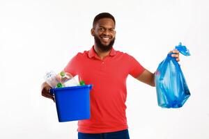 Cheerful Black Man Holding Blue Garbage Bag And Box With Recycle Symbol Plastic Litter Posing Over White Background In Studio. Keep Environment Clean And Sort Your Waste Concept photo