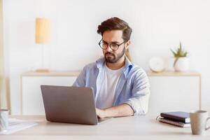 Telecommuting Concept. Focused Middle Eastern Man Using Laptop , Sitting At Desk In Home Office, Handsome Arab Freelancer In Eyeglasses Typing On Keyboard And Looking At Computer Screen, Free Space photo