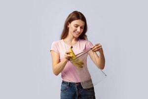 Zero Waste Shopping. Happy Millennial Woman Putting Bananas Into Reusable String Mesh Bag Instead Of Using Polythene Packet, Smiling Eco-Friendly Lady Standing On Gray Studio Background, Copy Space photo