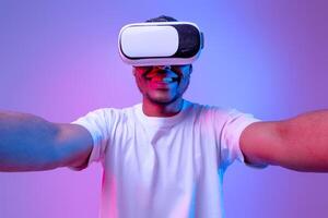 Virtual Entertainment Concept. Closeup Shot Of Excited Black Guy In VR Glasses Headset Extending Hands At Camera, Cheerful African American Man Standing In Neon Lighting Over Purple Background photo
