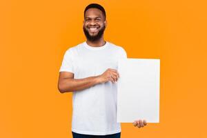 Cheerful African Man Showing Blank Paper Poster For Your Text Smiling To Camera Standing Over Yellow Studio Background. Black Guy Advertising Text Holding White Board. Mockup photo