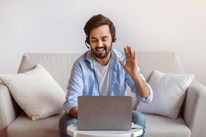 Friendly Eastern Man Making Call With Laptop At Home, Waving Hand At Web Camera, Greeting Somebody. Young Arab Guy Enjoying Online Communication While Sitting On Couch In Living Room, Copy Space photo