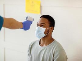 Young black man having his temperature measured by infrared thermometer at clinic before getting covid-19 vaccine injection. Population immunization and health service concept photo