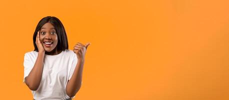 Amazed african american woman with beautiful hair pointing with thumb up at empty space, touching her face in surprised gesture and opening mouth, orange studio background, panorama photo
