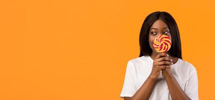 Shy black woman covering her mouth with candy on stick and looking at copy space for advert, orange studio background. Playful african american young lady having fun, holding sweets, panorama photo