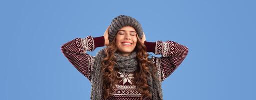 Portrait of lovely female model posing in warm knitted clothes over blue studio background, panorama. Gorgeous millennial lady wearing fashionable beanie hat, scarf and sweater with ornament photo