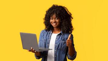 Happy african american young woman holding newest laptop and showing thumb up, yellow studio background, panorama with copy space. Cheerful black lady using notebook, shopping online or websurfing photo