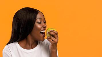 Healthy young black lady eating fresh green apple over orange studio background, panorama with copy space. Closeup of cheerful african american woman having sneak, enjoying juicy apple photo