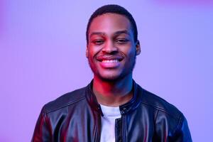 Portraif Of Happy Black Guy Posing In Leather Jacket Under Neon Light, Handsome Millennial African American Man Looking And Smiling At Camera While Standing Over Purple Violet Background, Copy Space photo
