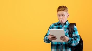 Amazed ginger boy schooler with backpack using digital tablet, looking at pad screen with shocked gace expression, emotional kid using educational application or playing cute mobile game, free space photo