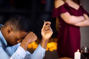 Desperate Black Man Sitting At Table With Ring In Hand After Girlfriend Refused His Proposal And Not Accepting Engagement Offer. Young African American Guy Feeling Lonely And Depressed, Cropped Image photo
