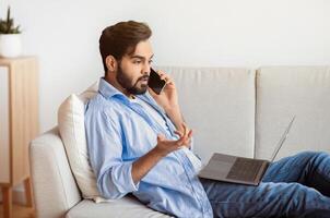 Arab Man Having Problem While Working With Laptop And Cellphone At Home, Eastern Freelancer Guy Suffering Trouble With Internet Connection, Speaking With Customer Service Manager, Sitting On Couch photo