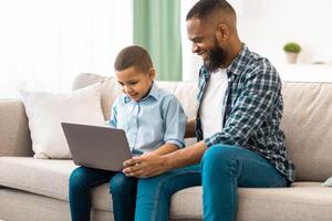 African Father And Son Using Laptop Browsing Internet Together Sitting On Couch At Home. Weekend With Dad. Little Boy And His Dad Watching Movie On Computer Online. Side View photo