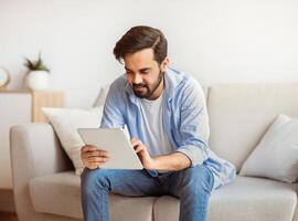 Handsome Eastern Guy Using Digital Tablet While Relaxing On Couch At Home, Millennial Arab Man Browsing New App, Working Online Or Making Shopping In Internet While Sitting In Living Room, Free Space photo