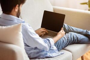 Unrecognizable Eastern Man Using Laptop Computer With Blank Black Screen While Sitting On Couch At Home, Working Online Or Browsing Internet Website, Over Shoulder View, Mockup Image With Copy Space photo