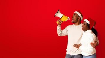 Christmas Announcement. Happy Black Couple In Santa Hats Shouting With Megaphone Loudspeaker At Copy Space On Orange Background, Smiling African Spouses Greeting With New Year And Xmas Holidays photo
