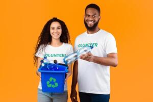 Two Cheerful Volunteers Holding Box With Recycling Symbol Full Of Wasted Plastic Bottles Standing On Yellow Background In Studio. Garbage Disposal, Sort And Recycle Your Waste Concept photo