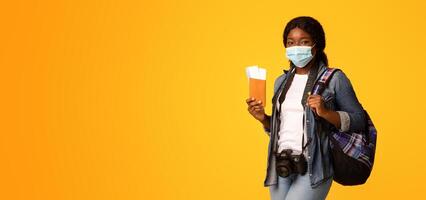 Travel During Pandemic. Young african tourist woman in medical face mask holding passport with tickets and photo camera, ready for vacation trip, standing isolated over yellow background, copy space