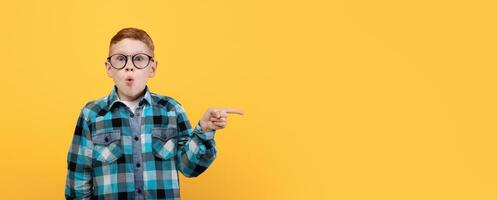 Amazed kid with glasses showing shocking advertisement, aiming at copy space for text or advertisement. Surprised redhead schoolboy pointing at empty space over yellow studio background, panorama photo