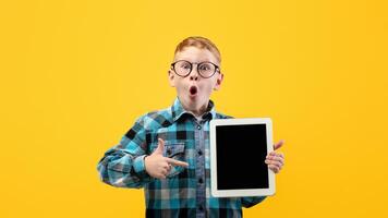 Amazed schoolboy in glasses pointing at empty digital tablet screen, recommending cool educational or entertaining website or application, mockup, yellow studio background, copy space photo