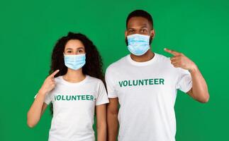 Female And Male Volunteers Pointing Fingers At Protective Masks On Their Faces Posing Over Green Background. Studio Shot. Corona Virus Covid-19 Protection, New Rules Concept photo