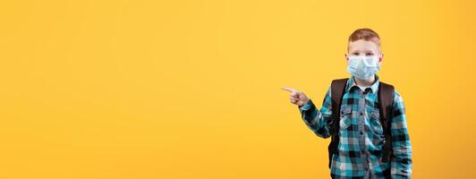 Schoolboy in protective face mask pointing at copy space over yellow studio background. Redhead schoolboy with backpack wearing face mask, using protection against coronavirus while going to school photo