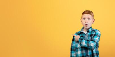 Amazed red-haired school boy in plaid shirt aiming at copy space over yellow studio background. Shocked ginger kid pointing at empty space, showing exciting advert for children photo