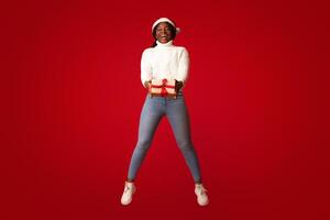 Cheerful black woman in santa hat jumping in air with christmas present in hands over orange background, excited african american lady holding wrapped present box and smiling, enjoying xmas holidays photo