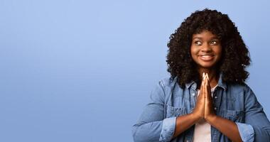 Inspired young black woman showing clasped hands in pleading gesture, pretty please on yellow studio background, looking at copy space. Religious african lady praying, having hope for better future photo