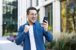 An excited man outside is looking at his phone, celebrating good news. He smiles broadly and raises his fist in joy, demonstrating his accomplishment. photo