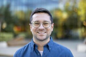 A portrait of a smiling man wearing glasses outdoors. He's dressed in a blue button-down shirt. A warm, inviting expression. photo