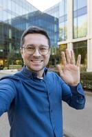 A man waves at the camera, smiling broadly, with a modern building in the background. The man wears glasses and a blue shirt. photo