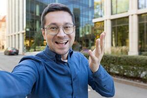 A man takes a selfie with a bright smile, gesturing toward the camera in front of a modern office building. photo