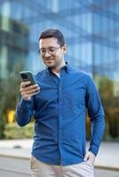 A man in glasses is using a smartphone outside in front of a modern office building, he seems pleased. photo