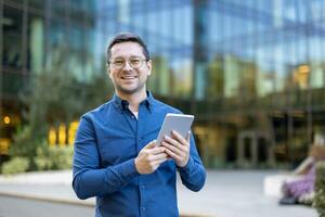 A smiling businessman in glasses holds a tablet outdoors in front of a modern building. It suggests business and technology. photo