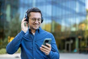 A man outside enjoys music through headphones, looking at his smartphone with a happy expression. The urban backdrop suggests a modern lifestyle. photo