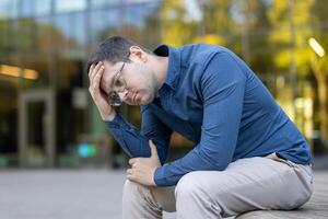 A man with glasses sits outside, looking down with a hand on his head, appearing stressed and worried, captured in daylight. photo
