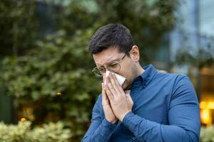 A man in glasses outside, blowing his nose with a tissue, suggesting allergies or illness. photo