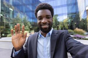 An attractive man in a business suit is waving in front of a modern building. Smiling directly at the camera, outdoors. photo