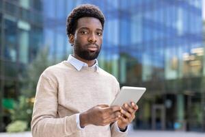 An african american man in a light sweater, holding a tablet, posing in front of an office building, looking at the camera. photo
