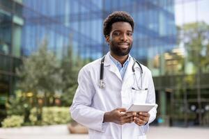 A smiling doctor in a white coat holds a tablet computer, appearing confident and professional in an outdoor setting with a building in the background. photo
