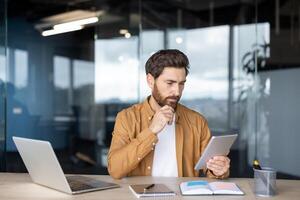 A thoughtful man with a beard is looking at a tablet in an office setting, possibly analyzing data or working on a project. photo