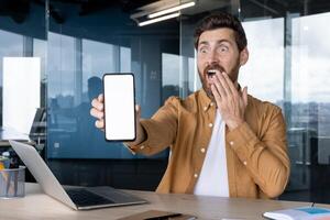 A man in an office is surprised while holding a blank smartphone screen, ideal for showcasing app displays or technology concepts. photo