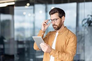 A focused businessman reviews a tablet while adjusting his glasses in a modern office setting, suggesting productivity and technology. photo