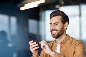 A smiling man in a button-down shirt looks at his phone, likely texting or browsing in a modern office setting. photo