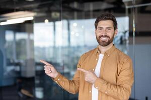 A smiling man in a smart casual outfit points towards an area with both hands, likely presenting something. photo