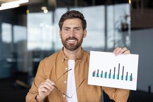 A man smiles during an online presentation, holding glasses and a chart with graphs, representing data analysis in a modern office environment photo