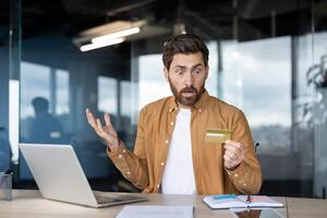 A man sits at a desk, looking shocked while holding a credit card near a laptop, possibly experiencing financial issues. photo
