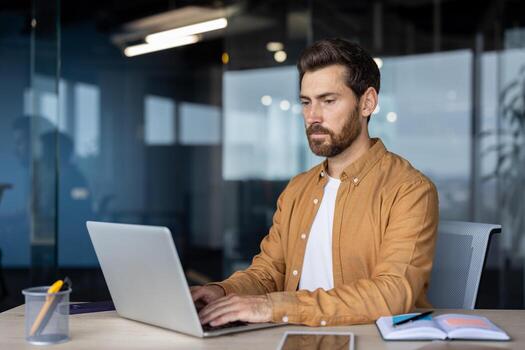 Serious man at work with laptop inside office, thinking businessman typing on keyboard, preparing electronic report, mature worker programmer, coding new software at workplace. photo